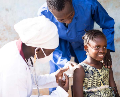 A smiling African girl getting a vaccine on her upper arm by a black female pediatrician assisted by a healthcare worker