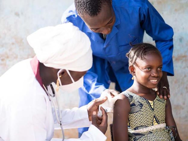A smiling African girl getting a vaccine on her upper arm by a black female pediatrician assisted by a healthcare worker